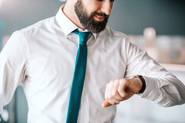 Ciérrese para arriba del hombre de negocios barbudo atractivo en camisa y ate mirando el reloj mientras que se coloca en oficina. Es hora de inspirar.