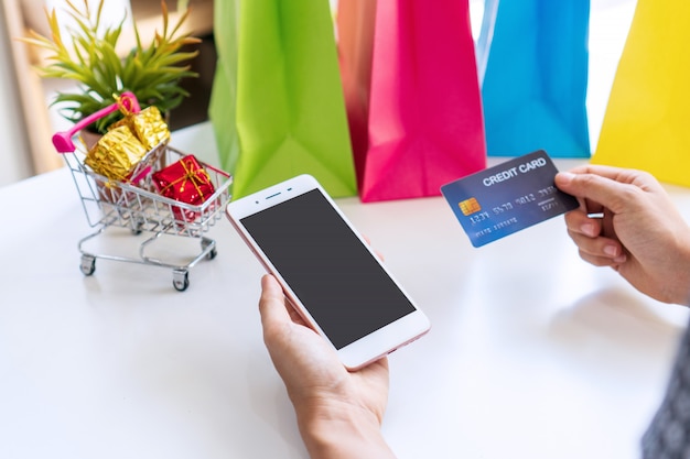 Ciérrese para arriba de la mano de la mujer que sostiene la tarjeta de crédito mientras que usa smartphone con las cajas de regalo miniatura en carretilla y bolsos coloridos en el fondo blanco.