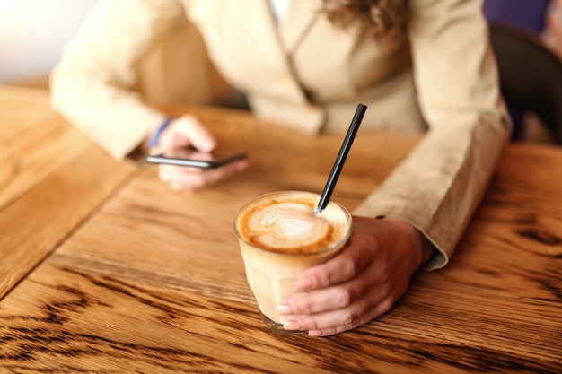 Ciérrese para arriba de mujer caucásica en chaqueta usando el teléfono elegante y bebiendo café mientras que se sienta en cafetería. Enfoque selectivo en mano con café.