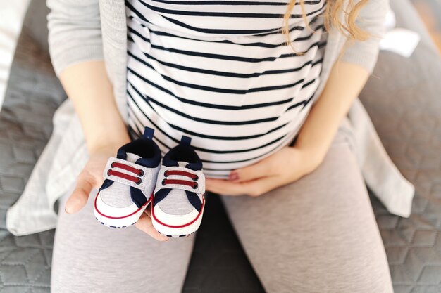 Ciérrese para arriba de la mujer caucásica embarazada que sostiene los zapatos de bebé y que toca el vientre.