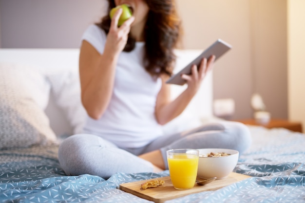 Ciérrese para arriba de la mujer envejecida media atractiva sonriente que se sienta en la cama con una manzana y que mira en una tableta.