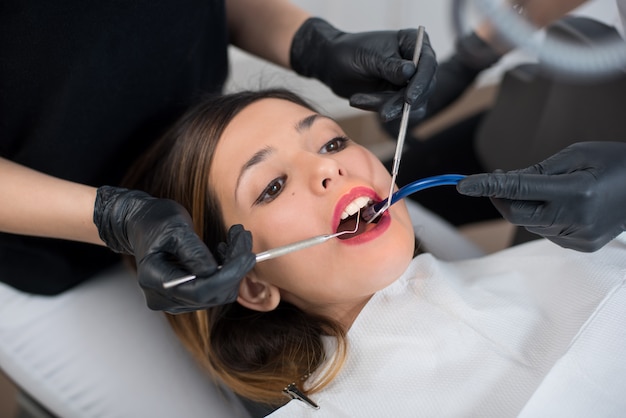 Ciérrese para arriba de la mujer joven hermosa que tiene chequeo dental. Dentista examinando los dientes de un paciente, sosteniendo herramientas dentales - espejo y sonda. Odontología.