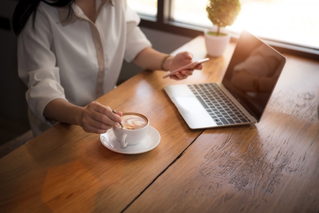 Ciérrese para arriba de la mujer de negocios que trabaja con el ordenador portátil y que bebe el café en oficina