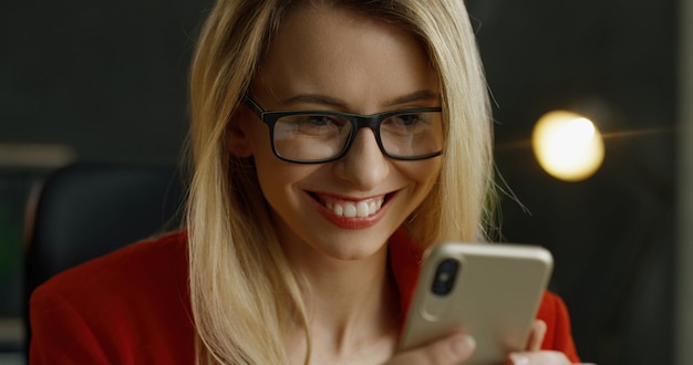 Ciérrese para arriba de smartphone en manos de la empresaria rubia. Feliz mensaje de texto femenino y escribiendo en el teléfono celular en el gabinete.