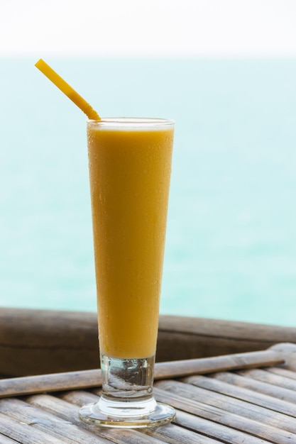 Ciérrese para arriba en el vaso largo del jugo de mango en la tabla de bambú por el mar. Bebida fría, concepto de sed