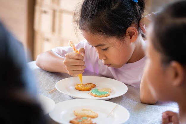 Ciérrese encima de la muchacha asiática del niño que exprime la crema de hielo para decorar las galletas con un amigo con la diversión. Concepto de arte y artesanía infantil hecho en casa de bricolaje.