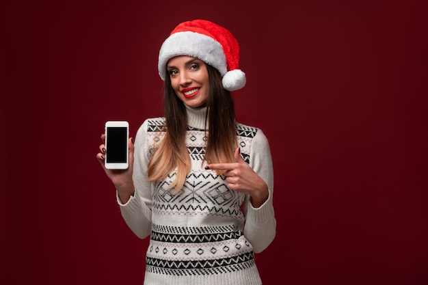 Ciérrese encima de la mujer caucásica beautifiul del retrato en el sombrero rojo de Santa en la pared roja. Concepto de Navidad año nuevo. Linda mujer dientes sonriendo emociones positivas con espacio de copia libre