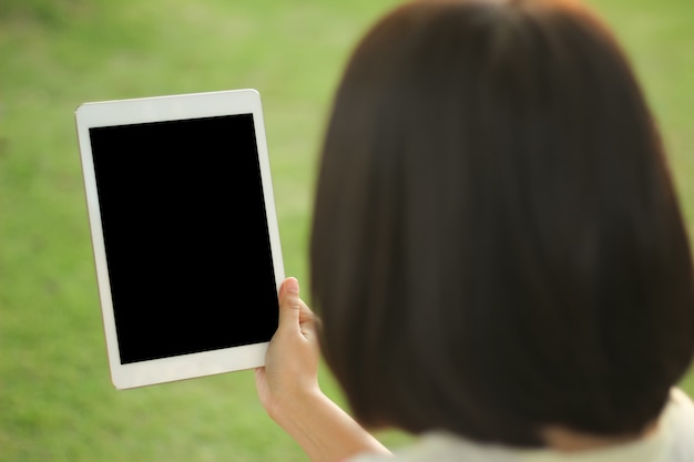 Ciérrese encima de la mujer que sostiene y que usa la tableta mientras que se sienta en al aire libre, trabajando en parque