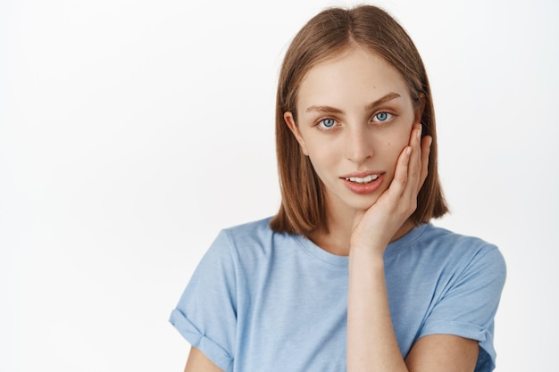 Ciérrese encima del retrato de la cara de la mujer joven hermosa, del pelo corto y de los ojos azules, tocando la piel brillante natural limpia sin acné, sin maquillaje, sonriendo tiernamente en la pared blanca del frente.