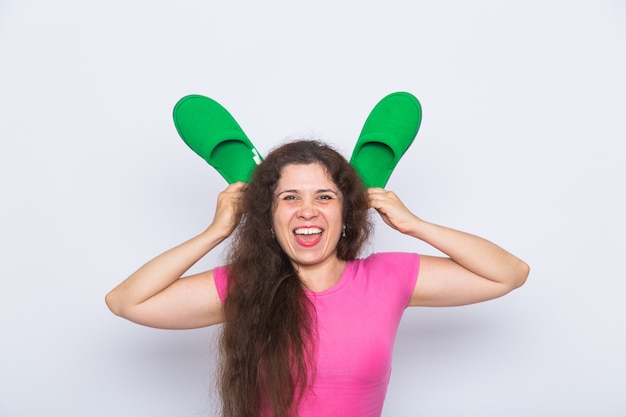 Ciérrese encima del retrato divertido loco de la mujer, haciendo muecas, imitando orejas de conejo con zapatillas sobre fondo blanco