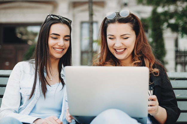 Ciérrese encima del retrato de dos amigas encantadoras que se divierten mientras está sentado en un banco riendo y mirando a una computadora portátil afuera en el parque.