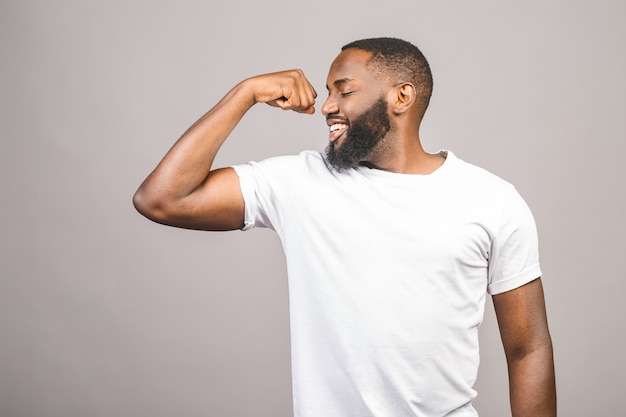 Ciérrese encima del retrato de un hombre afroamericano joven feliz que dobla el músculo del bíceps aislado contra fondo gris.