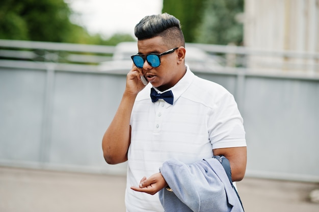 Ciérrese encima del retrato del hombre árabe elegante en las gafas de sol y la corbata de lazo presentadas al aire libre y hablando en el teléfono móvil. Hombre modelo árabe.