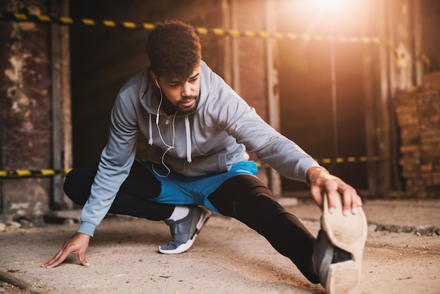 Ciérrese encima del retrato del hombre atlético atractivo joven afroamericano activo que hace entrenamiento completo del estiramiento de la pierna dentro del lugar abandonado.