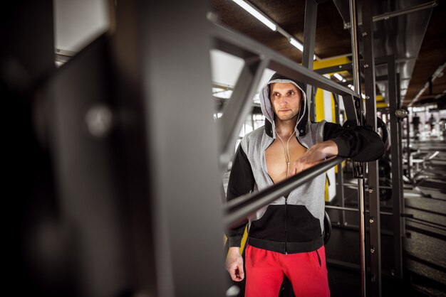 Ciérrese encima del retrato del hombre encapuchado muscular enfocado y motivado que se coloca con los auriculares en el gimnasio moderno.