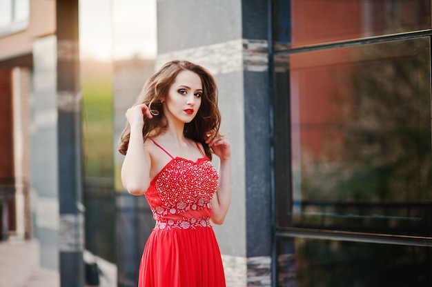 Ciérrese encima del retrato de la muchacha de moda en el vestido de noche rojo que plantea la ventana del espejo del fondo del edificio moderno