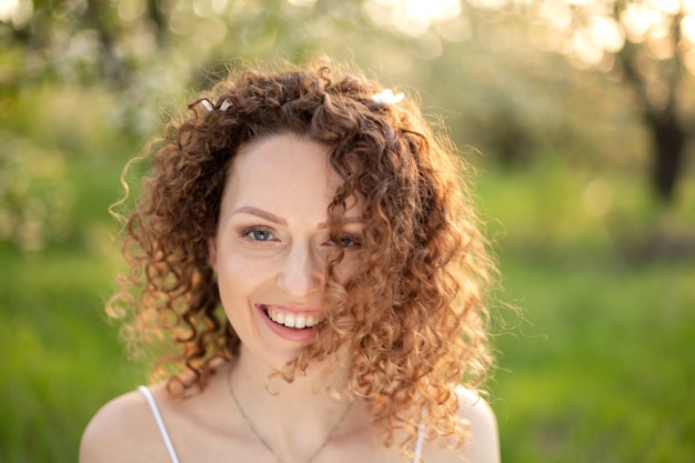 Ciérrese encima del retrato de la mujer atractiva sonriente de los jóvenes con el pelo rizado en parque floreciente verde de la primavera. Emociones puras