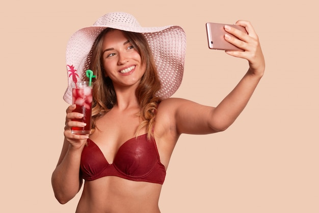 Ciérrese encima del retrato de la mujer caucásica atractiva que toma el selfie mientras que sostiene el cóctel fresco disponible. Hermosa mujer joven en traje de baño rojo y sombrero de paja posando de nuevo pared beige estudio.