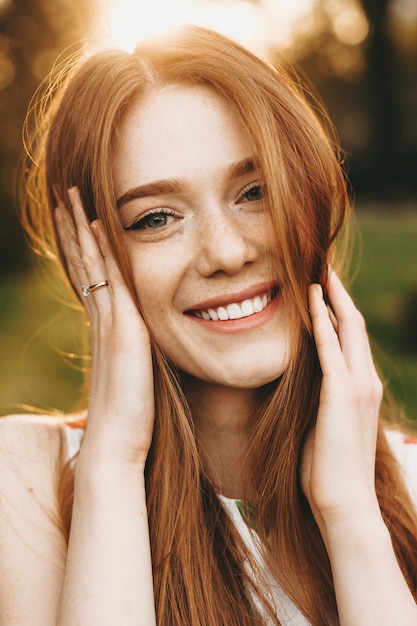 Ciérrese encima del retrato de una mujer joven alegre con el pelo largo rojo y las pecas mirando directamente sonriendo mientras sostiene ambas manos en su cabello contra la puesta del sol.