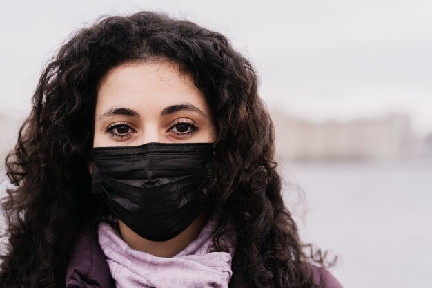 Ciérrese encima del retrato de una mujer morena joven con los ojos marrones grandes en la máscara de la medicina en su cara que piensa en virus.