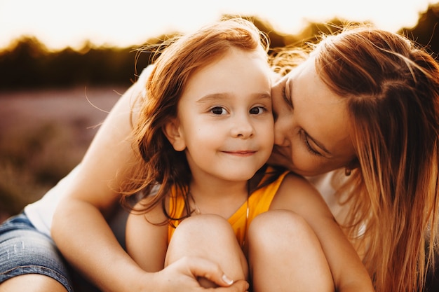 Ciérrese encima del retrato de una niña asombrosa que mira la cámara sonriendo mientras su madre la abraza y la besa contra la puesta del sol.