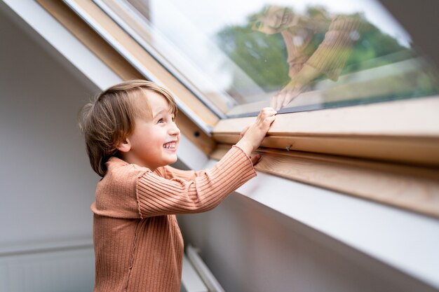 Ciérrese encima del retrato de un niño preescolar pequeño que mira la ventana y que ríe. Niño sonriente feliz pasa tiempo solo en casa.