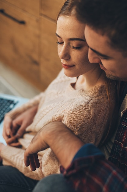 Ciérrese encima del retrato de una pareja joven encantadora que se abraza mientras que el hombre está abrazando a su novia desde atrás sentada en el suelo con los ojos cerrados.