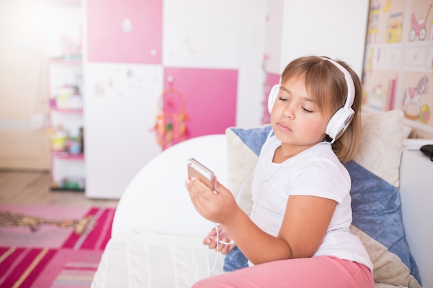 Ciérrese encima del retrato de la pequeña muchacha adorable caucásica que escucha la música en auriculares en el dormitorio acogedor que sonríe y que se relaja.