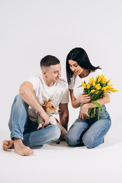 Ciérrese encima del retrato que los pares jovenes se sientan y que abrazan, sosteniendo las flores y el perro amarillos en estudio en el fondo blanco. Pareja abrazada con expresión amorosa de ensueño. Familia amorosa. Celebrando el día de la mujer.