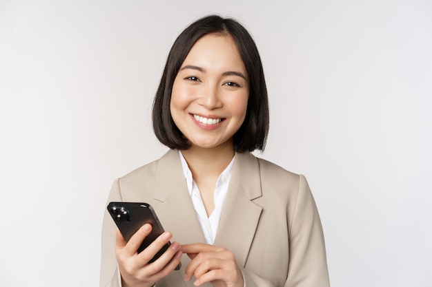 Ciérrese encima del retrato de la señora corporativa de la mujer coreana en traje que usa el teléfono móvil y que sonríe que sostiene el teléfono inteligente que se coloca sobre el fondo blanco