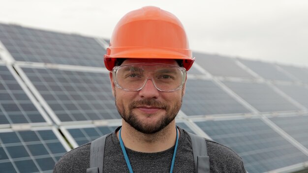 Ciérrese encima del retrato del trabajador eléctrico masculino en el casco protector que se coloca cerca del panel solar. Producción de energía limpia. Energía verde. Granja solar ecológica.