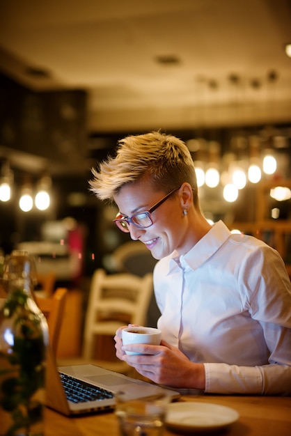 Ciérrese encima de vista lateral de la mujer blogger sonriente confiada elegante del pelo corto que sostiene una taza de café mientras que se sienta en un café y trabaja en una computadora portátil en la noche.