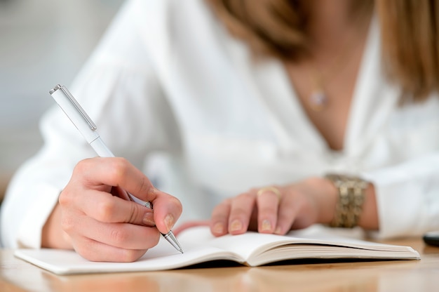 Ciérrese encima de la vista de la mano de la mujer usando la escritura de la pluma en el cuaderno mientras que se sienta en la tabla de madera.