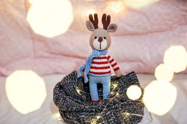 Ciervo de navidad. Ciérrese encima de los ciervos del amigurumi del juguete en suéter rayado y bufanda azul elegante con las luces de Navidad alrededor.