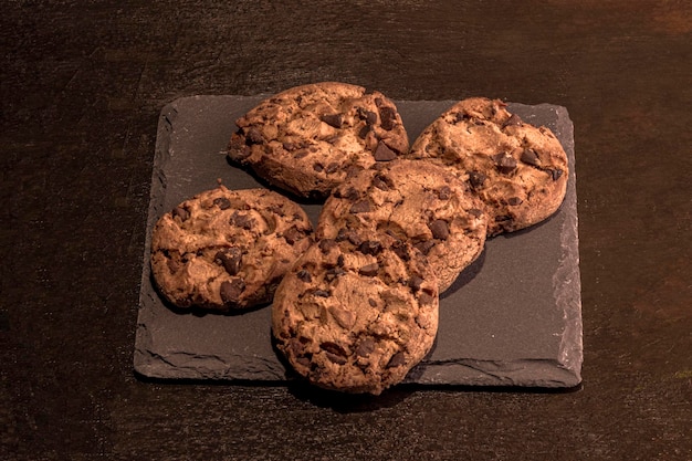 Cinco biscoitos com pedaços de chocolate em uma laje de ardósia em um fundo marrom pintado à mão.