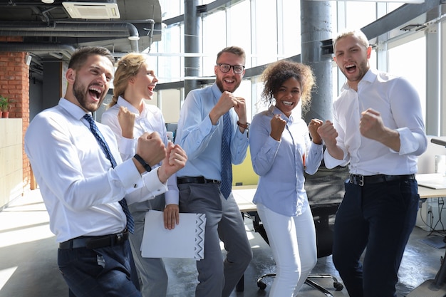 Cinco felices empresarios modernos mantienen los brazos en alto y expresan alegría mientras están parados en una oficina grande.