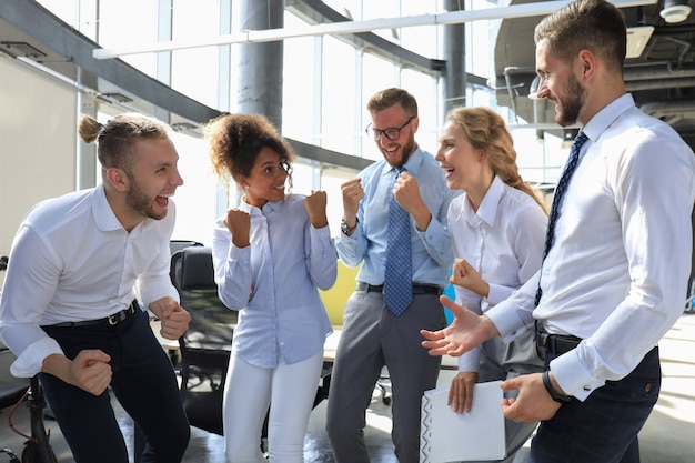 Cinco felizes empresários modernos estão mantendo os braços levantados e expressando alegria enquanto estão em um grande escritório.