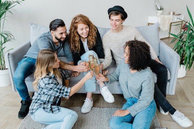 Cinco jóvenes amigos alegres en ropa casual tintineando con flautas de champán mientras celebran las vacaciones en casa