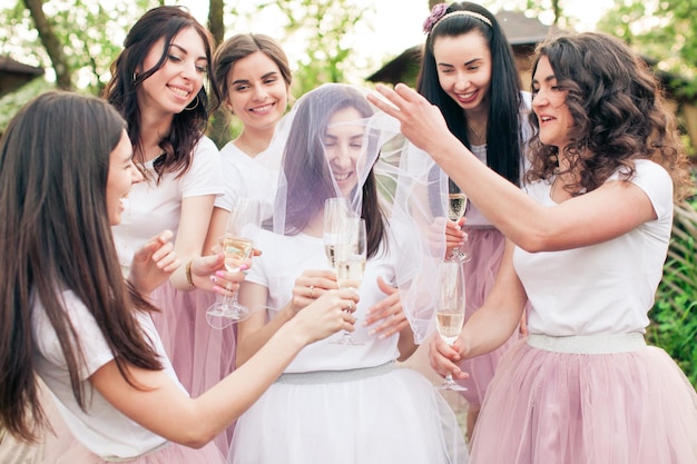 Cinco novias felices novias de pie alrededor de la joven novia con velo blanco en la cara. Chicas con faldas de tul rosa claro y camisetas blancas, con copas de champán en las manos