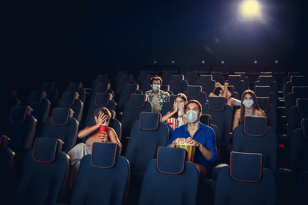 Cine, sala de cine durante la cuarentena. Reglas de seguridad para la pandemia de coronavirus, distancia social durante la visualización de películas. Hombres y mujeres con mascarilla protectora sentados en filas de auditorio, comiendo po