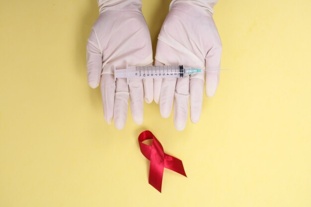 Foto cinta roja con guantes médicos y jeringa contra el vih aislado sobre fondo amarillo