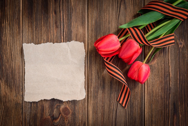 Cinta de San Jorge, tulipanes rojos y tarjeta de felicitación de papel sobre fondo de madera. Día de la victoria o día del defensor de la Patria.