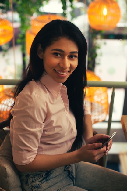 Cintura para arriba de una bella dama en ropa casual sentada con un moderno teléfono inteligente en sus manos y sonriendo a la cámara. Lámparas en el fondo