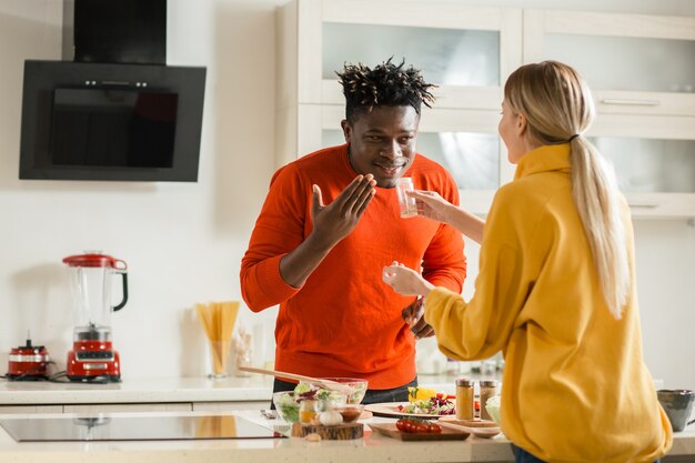 Cintura para arriba de la dama de pelo largo de pie en la cocina frente a su alegre novio y dándole especias