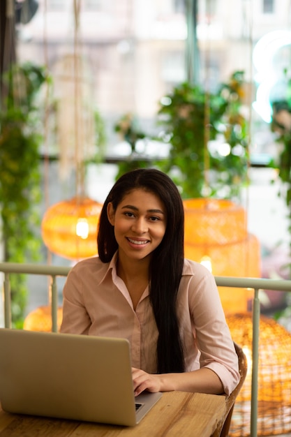 Cintura para arriba de una joven complacida trabajando en su computadora portátil y sonriendo a la cámara