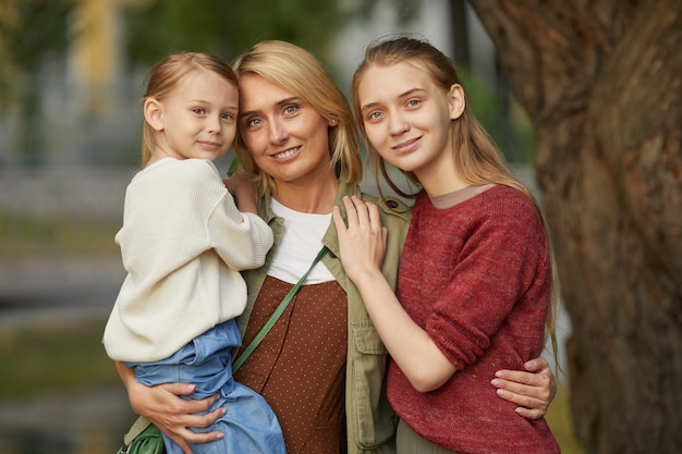 Cintura para arriba madre adulta moderna con dos hijas posando juntas y sonriendo felizmente mientras está de pie junto al árbol al aire libre disfrutando de tiempo en familia en el parque.