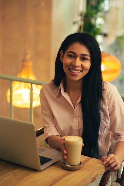 Cintura para arriba de una mujer joven de pelo largo sentada en la mesa de café con una computadora portátil moderna frente a ella y sonriendo mientras toca un vaso de café con leche