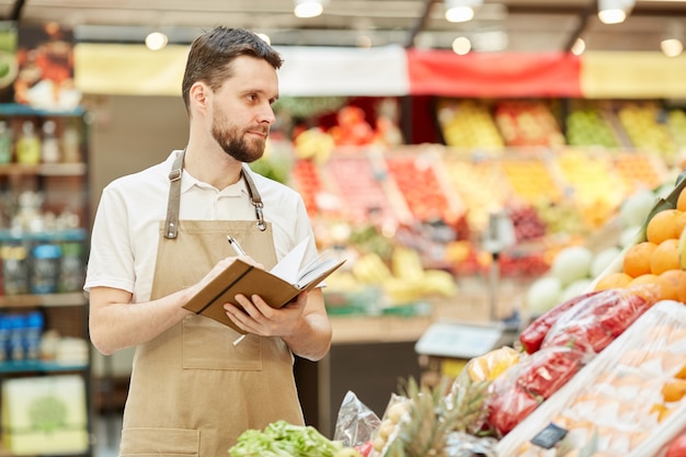 Cintura para arriba retrato de agricultor barbudo sosteniendo portátil mientras vende frutas y verduras frescas en el mercado stand