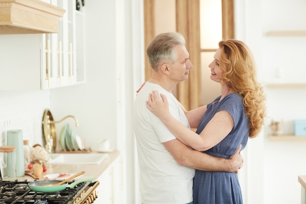 Cintura para arriba retrato de amorosa pareja madura abrazándose y mirando el uno al otro mientras está de pie en el interior de la cocina blanca en casa, espacio de copia