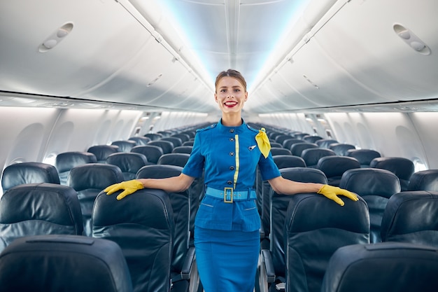 Cintura para arriba retrato de asistente de vuelo de mujer sonriente feliz en uniforme azul con guantes amarillos mientras ella está poniendo las manos en las acogedoras sillas de pasajeros